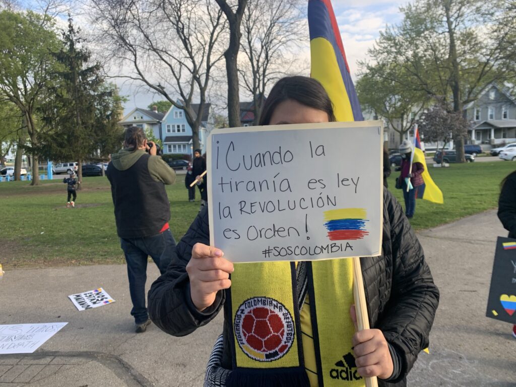 Protestor wearing a Colombian scarf and holding a Colombian flag holds up a sign in front of their face that says "Cuando la tiranía es ley, la revolución es orden." Translation: "When tyranny is law, revolution is order." 