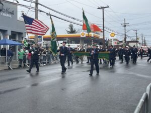 Men dressed in military uniforms are carrying three flags: an American flag, an Irish flag, and a flag commemorating St. Patrick’s Day as they march in the parade.