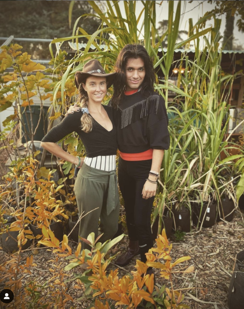 Gillian and Julian. both smiling, are standing next to each other in front of large plants.