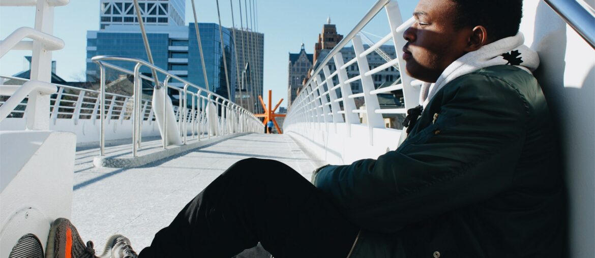 Man sitting on bridge over Milwaukee river, with The Calling sculpture in the distance