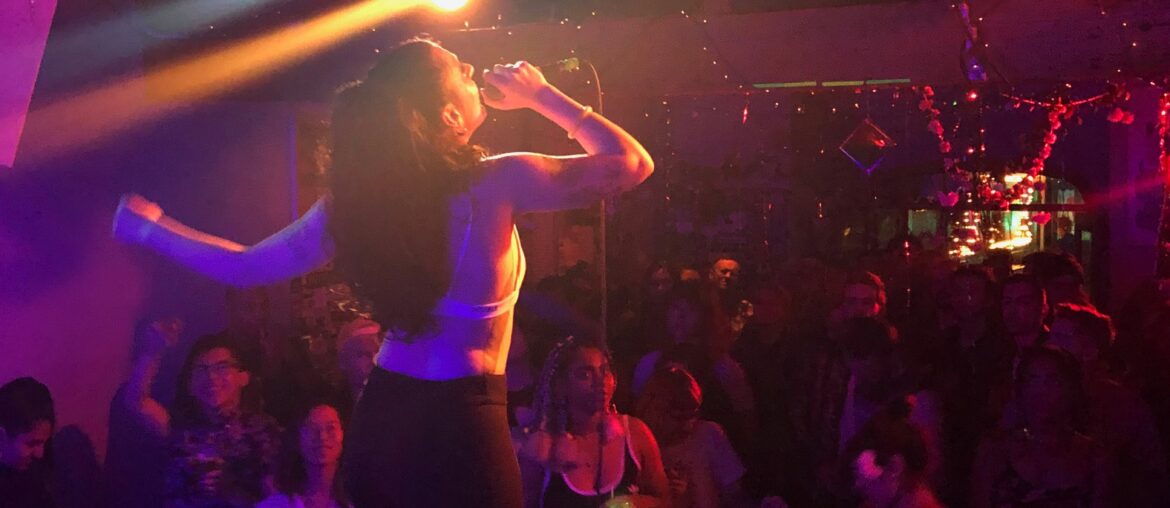 A woman on the stage in front of a small crowd in a dim nightclub