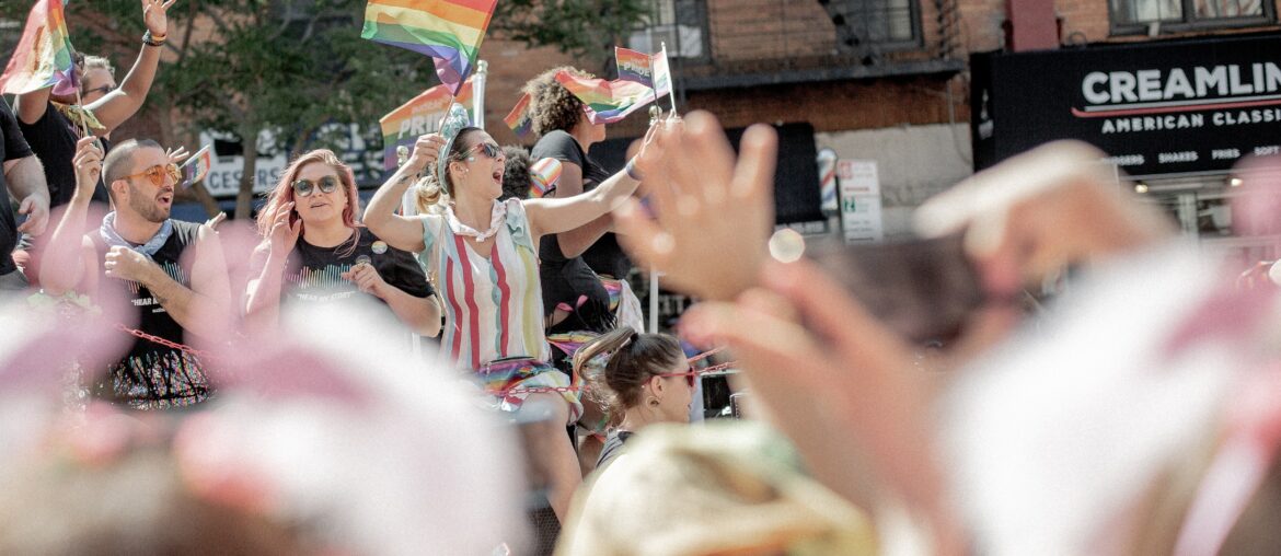 People celebrating on top of a bus, waving rainbow flags with the crowd in the blurred foreground