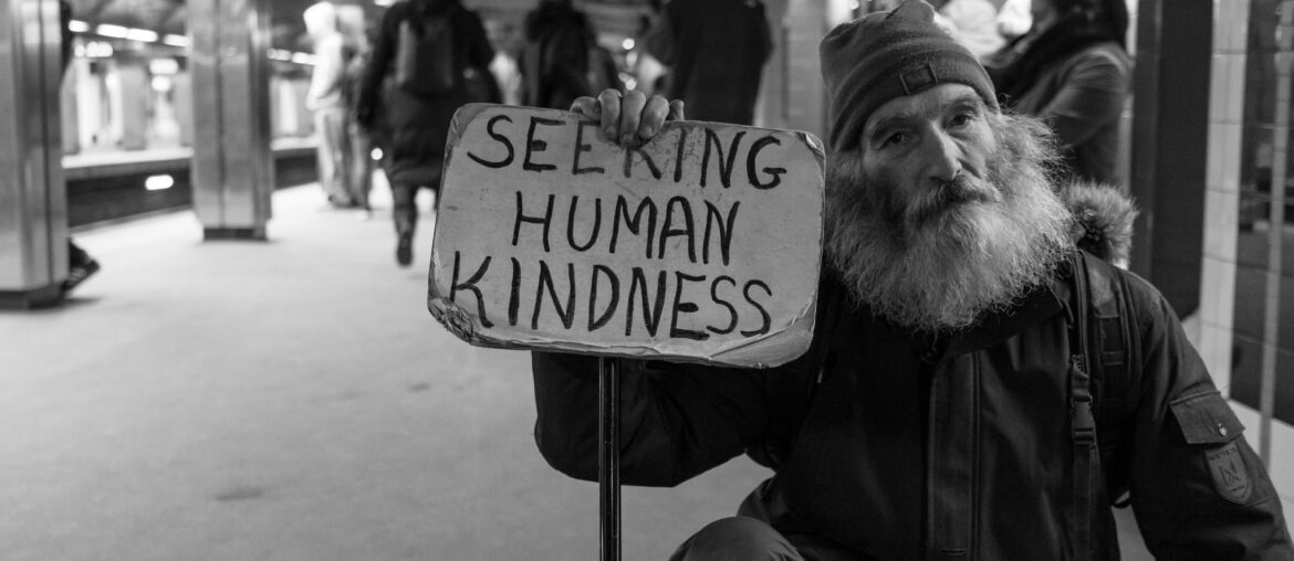 man with white beard hold up sign say "seeking human kindness" in crowded subway