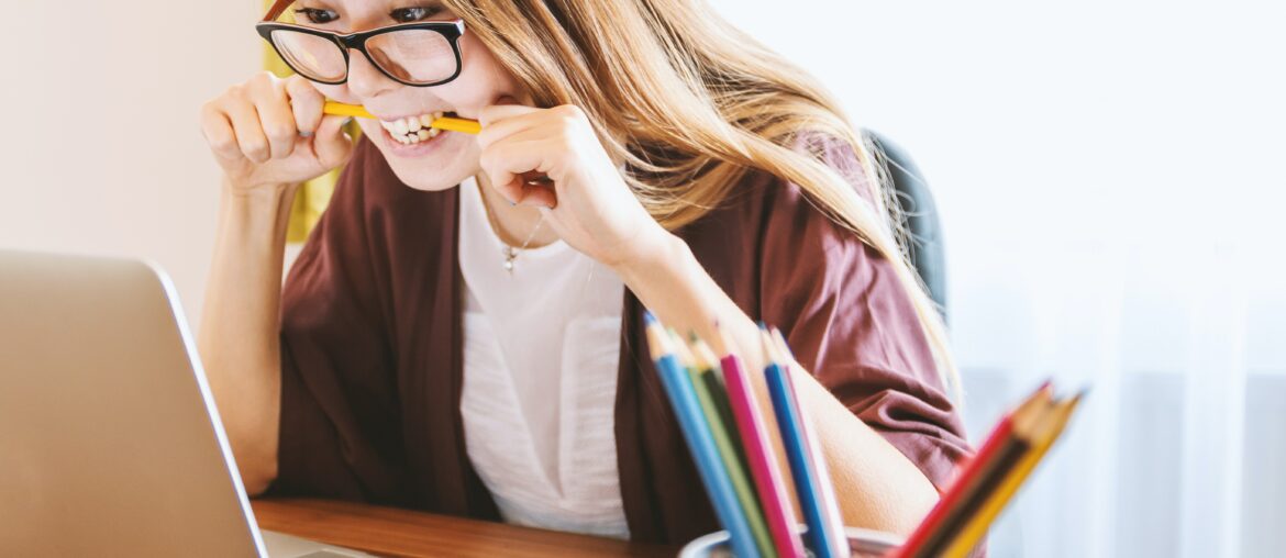 woman with glasses biting on to pencil while looking at laptop