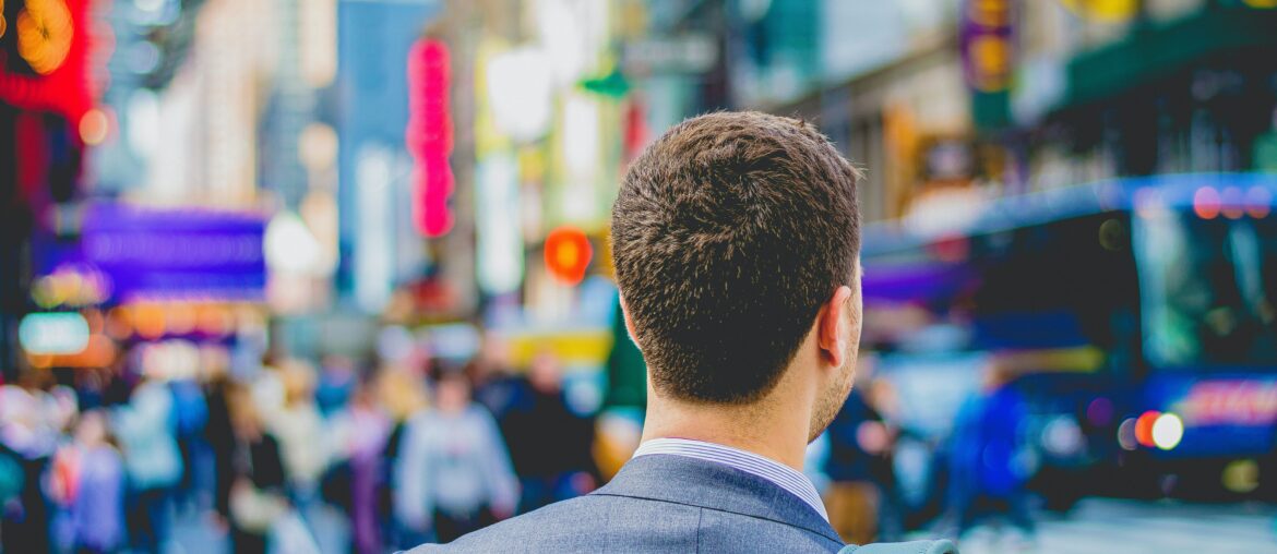 Man wearing suit has a backpack over his shoulder as he looks on at a bustling Times Square in New York City
