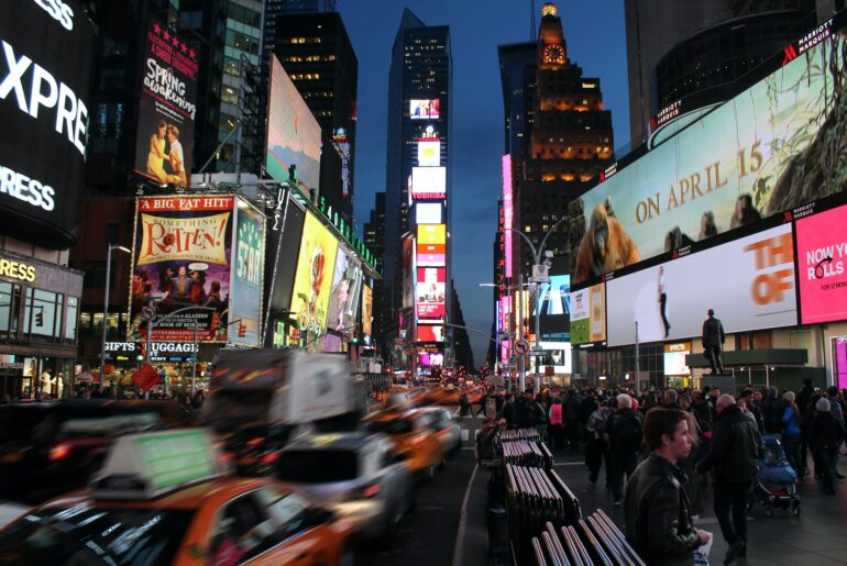 Bright lights in Times Square, New York at night. Cars and traffic blurred in the foreground
