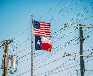American USA Flag and Texas State Flag flying together in front of power lines. The state of Texas operates its electrical grid through a deregulated market, overseen by the Electric Reliability Council of Texas (ERCOT), who coordinates the flow of electricity and ensures grid reliability.